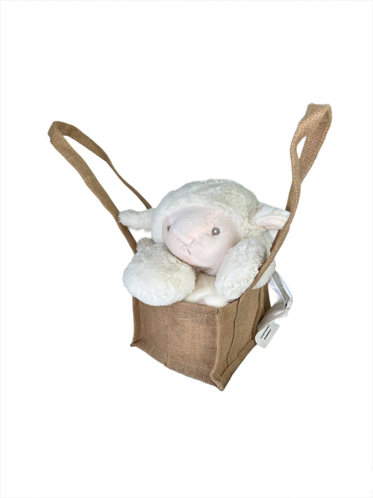 White-marshmellow coloured fluffy plush lamb with a pale peach face, head resting adorably on its paws, sitting propped up in a natural, brown hessian jute bag